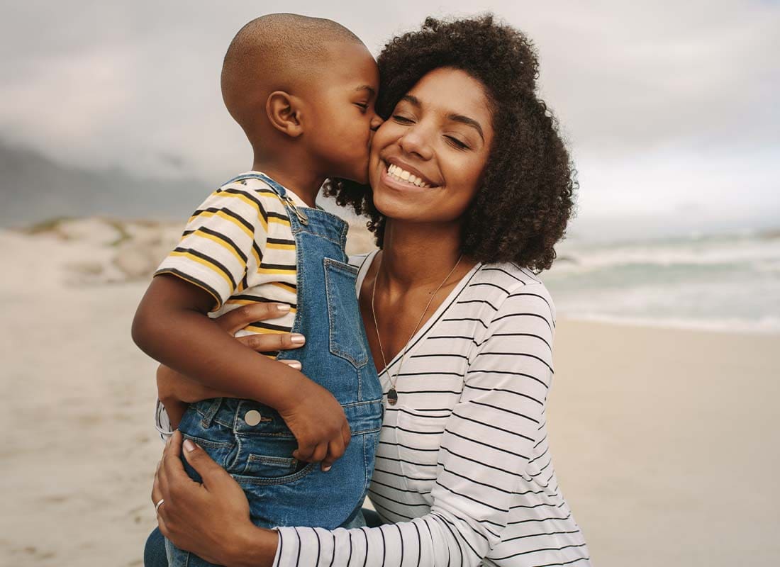 Life Insurance - Young Son Kissing His Smiling Mother While Being Held at the Beach on a Cloudy Day